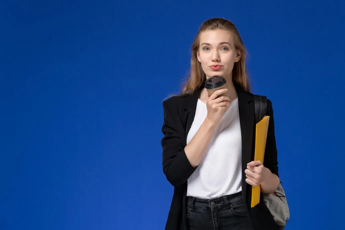 Frontansicht einer Studentin in schwarzer Jacke mit Rucksack, die eine gelbe Mappe und einen Kaffee hält, vor einer blauen Wand; Schule, College, Universität, Unterricht.